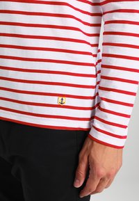 Maglietta a maniche lunghe a righe rosse e bianche con un piccolo logo di ancora gialla sul lato sinistro, abbinata a un tessuto nero.