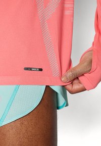 Koraalkleurig longsleeve shirt met meshpatronen en een "ACTIBREEZE" label, gecombineerd met lichtblauwe shorts met een gestructureerd design en een rond afgewerkte zoom.