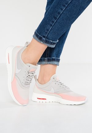 Stopy w niebieskich podwiniętych dżinsach i lekkich szarych sneakersach z różowymi akcentami oraz białymi podeszwami, ustawione na białej podłodze.