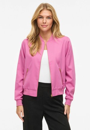 Femme portant une veste bomber rose zippée sur un haut blanc et un pantalon noir, debout devant un fond blanc uni.
