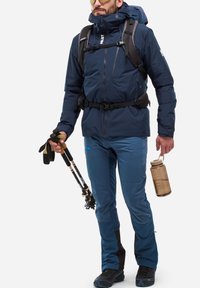 Navy waterproof jacket with a hood, paired with blue pants and black hiking boots. Holds trekking poles and a clear water bottle.