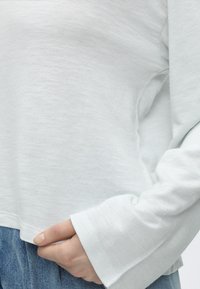 Άτομο που φοράει λευκό πουκάμισο με μακριά μανίκια και ανοίγματα για τον αντίχειρα, κρατώντας το τελείωμα του πουκάμισου πάνω από μπλε τζιν.