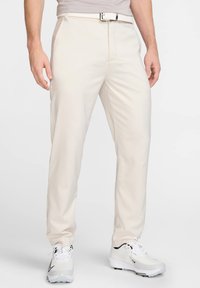 Pantalones beige de tela suave; diseño de pierna recta con trabillas para cinturón y un sutil pliegue frontal, usados con zapatos de golf blancos.