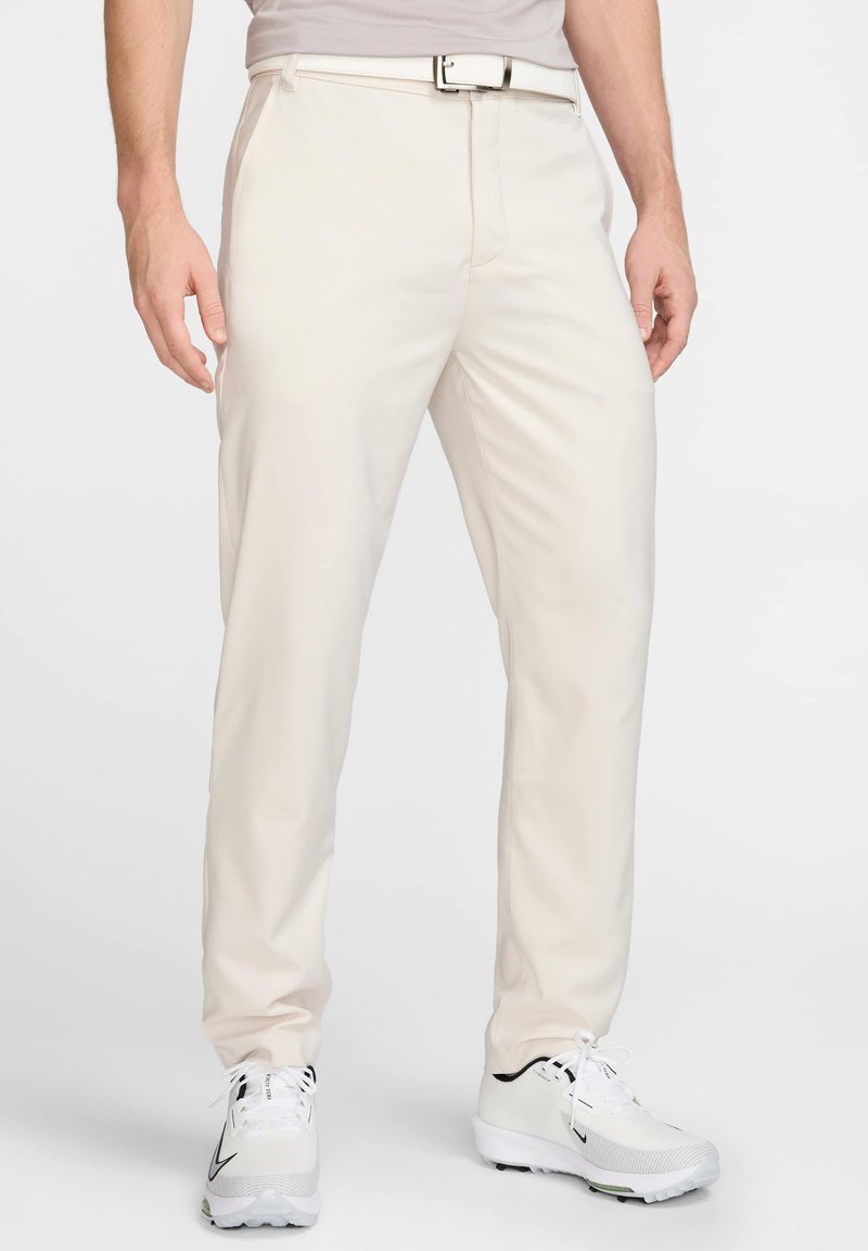 Pantalones beige de tela suave; diseño de pierna recta con trabillas para cinturón y un sutil pliegue frontal, usados con zapatos de golf blancos.
