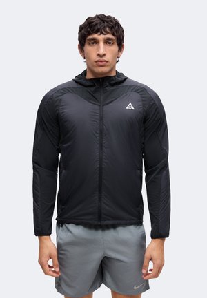 Homme portant une veste légère noire zippée avec capuche et un short de sport gris, debout devant un fond clair uni.