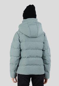 Chaqueta de plumas azul claro con capucha, tejido texturizado y secciones acolchadas en horizontal. Lleva puesta sobre pantalones negros y un gorro negro.