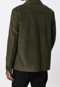 Oliwkowa kurtka z cordurowego materiału o fakturowej powierzchni, z guzikami przy mankietach oraz designem w okolicy pleców. Ma pionowe prążki i prosty krój.