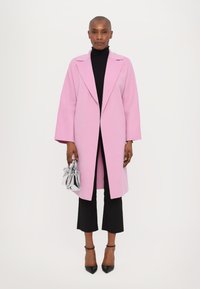 Palton din lână de culoare roz deschis, cu revere largi, purtat peste un pulover negru cu guler înalt. Modelul ține o geantă mică, strălucitoare, din argint. Pantaloni cropped negri și pantofi cu toc.