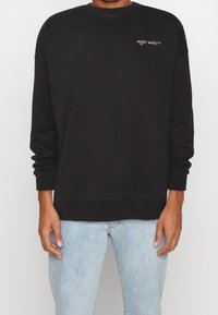 Sudadera negra con mangas largas, cuello redondo y texto blanco bordado. Combinada con jeans de mezclilla azul claro. Textura suave.