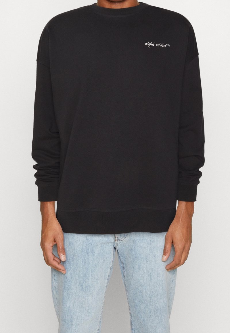 Sudadera negra con mangas largas, cuello redondo y texto blanco bordado. Combinada con jeans de mezclilla azul claro. Textura suave.