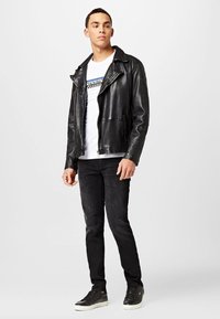 Chaqueta de motorista de cuero negro con cuello ancho, cremallera frontal y bolsillos laterales, usada sobre una camiseta gráfica blanca y unos jeans negros ajustados con zapatillas.