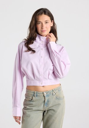 Jonge vrouw die een cropped, lichtpaarse gestreepte blouse met knopen en vervaagde blauwe spijkerbroek draagt, poseren met één hand bij haar hals.