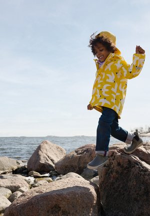 Gelbe Regenjacke mit Vogelmuster, graue wasserdichte Stiefel und blaue Jeans; Kind, das auf Steinen nahe einem Gewässer unter einem hellen Himmel läuft.