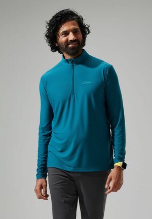Türkisfarbendes langärmeliges Zip-Up-Shirt aus leichtem Material, mit flachen Nähten und einem dezenten Logo auf der Brust. Kombiniert mit grauen Hosen.