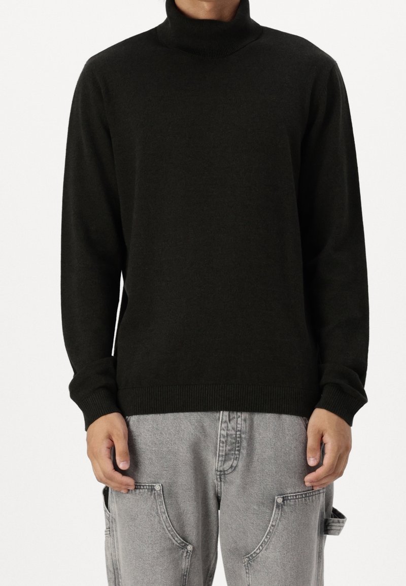 Mężczyzna w czarnym swetrze z wysokim kołnierzem i jasnoszarych spodniach cargo, stojący z rękami luźno wzdłuż ciała na jednolitym tle.