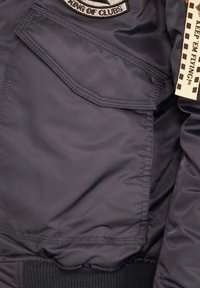 Schwarze Bomberjacke mit einer Reißverschlusstasche, gerippten Bündchen, glatter Textur und einem Etikett mit karierter Gestaltung. Gestickter Aufnäher auf der Brust.