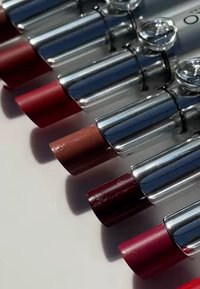 Colección de barras de labios en tonos variados, con tubos metálicos plateados y puntas de colores en tonos rojo, bayas y nude con acabados brillantes.