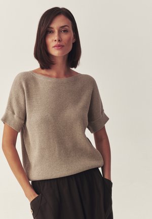 Kobieta o prostych brązowych włosach, ubrana w beżowy krótki sweter z krótkim rękawem i czarne spodnie, stojąca z rękami w kieszeniach na jednolitym tle.