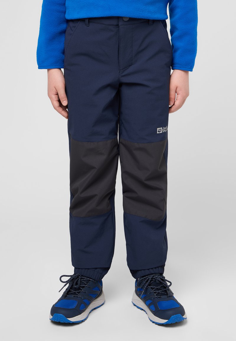 Pantaloni impermeabili blu navy con un pannello grigio scuro a contrasto, vita elastica e polsini regolabili, abbinati a scarpe da ginnastica blu.