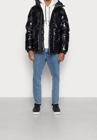 Giacca a vento nera lucida con cappuccio, dotata di due tasche frontali e chiusura con zip, indossata sopra un hoodie bianco e jeans blu con sneaker nere.