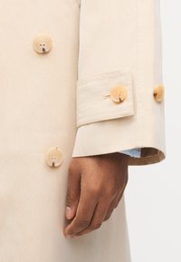 Χέρι που ακουμπά δίπλα στο μανίκι μιας μπεζ μπουφάν τύπου trench με μανσέτα με κουμπιά και εμφανή μεγάλα μπεζ κουμπιά στο μπροστινό μέρος της μπουφάν.