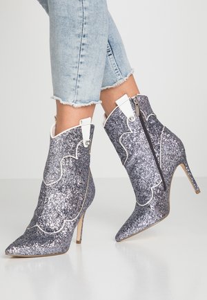 Kobieta nosząca srebrne, błyszczące botki na wysokim obcasie z białą lamówką i bocznymi zamkami, zestawione z jasnoniebieskimi, postrzępionymi obcisłymi dżinsami.