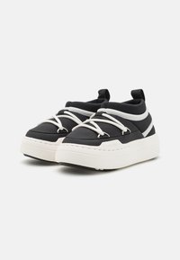 Sneaker bianche e nere con una spessa suola bianca, dotate di lacci elastici e di un colletto alla caviglia imbottito. Materiale sintetico liscio e design minimalista.