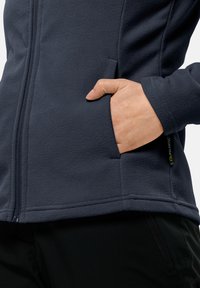 Giacca in pile di un blu scuro con zip frontale, tasche laterali e logo Polartec. La texture morbida e il design aderente migliorano il calore e il comfort.