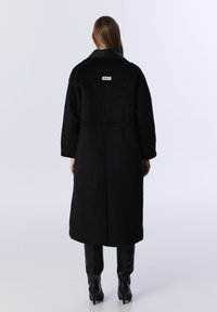 Schwarzer Wollmantel mit Lederkragen, langen Ärmeln und geradem Schnitt. Mit einem Logo auf der Rückseite und glatter Textur. Knöchelhoche Stiefel.