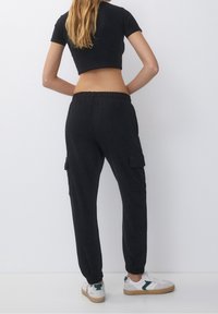 T-shirt noir court et pantalon cargo noir avec des poignets élastiques. Le pantalon comporte des poches latérales. Le modèle porte des baskets blanches avec des accents verts.