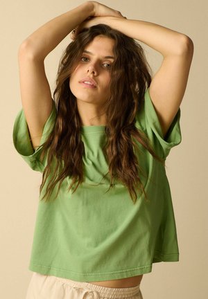 Junge Frau mit langen braunen Haaren trägt ein grünes kurzärmeliges Hemd und beige Hosen und hebt beide Arme über den Kopf vor einem neutralen Hintergrund.