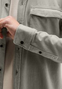 Helles graues Hemd aus weichem Stoff, mit einer Brusttasche, Knopfmanschetten und sichtbaren Nähten am Kragen und der Tasche.