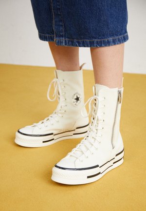 Hvide høje Converse-sneakers med sidelommer og sort stjerne-logo, båret med cropped blå jeansbukser på en gul gulv.