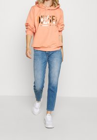 Sudadera con capucha color durazno y logo dorado, combinada con jeans rectos de color azul claro y zapatillas blancas. Un atuendo casual y cómodo.