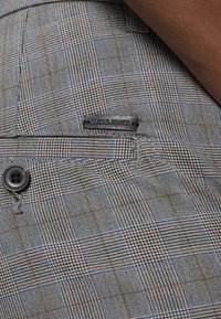 Tessuto a quadri grigio e blu con un accento marrone e una piccola etichetta nera. Include un bottone nero e dettagli con trama testurizzata.