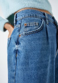 Jeans de denim azul claro con cintura alta, que cuentan con remaches de cobre, un desvanecimiento sutil y un diseño clásico de cinco bolsillos. Vista en detalle.