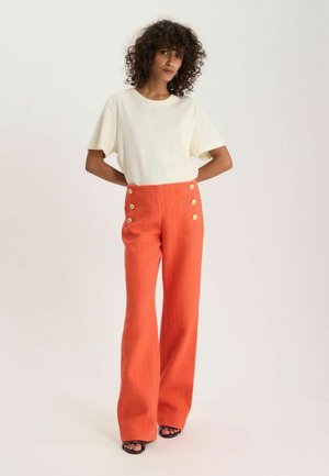 Femme aux cheveux bouclés portant un top blanc à manches courtes et un pantalon orange taille haute à jambes larges avec des boutons décoratifs, debout.