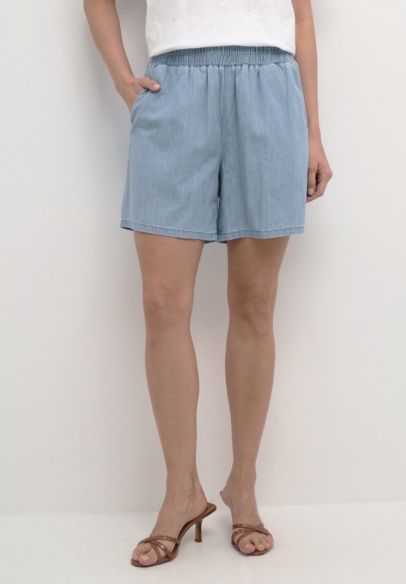 Vaaleansiniset farkkushortsit, joissa on joustava vyötärö, kaksi sivutaskua ja rento leikkaus, yhdistettynä ruskeisiin sandaaleihin, joissa on korkeat korot ja hihnat.