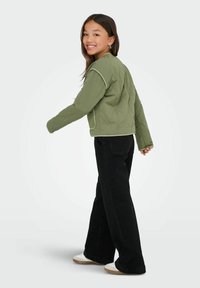 Olivgrön quiltad jacka med vit kant, som har en avslappnad passform. Buren med svarta vida byxor och vita sneakers, vänd något bort.