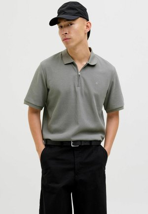 Polo de manga corta gris con un acabado texturizado, cuello con cremallera y un pequeño logo en el pecho, combinado con pantalones negros y una gorra negra.