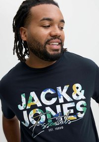 Crna pamučna majica s kratkim rukavima koja prikazuje odvažan, višebojan grafički tekst "JACK & JONES Essentials" s dizajnom s prskanjem. Okrugli izrez.
