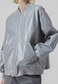 Veste bomber grise en matériau brillant et lisse. Comprend un col et des poignets côtelés, une fermeture éclair à l'avant et des poches latérales.
