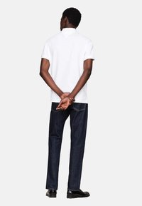 Camisa polo blanca de manga corta combinada con jeans oscuros de denim, diseño ajustado, textura suave y detalle de cuello clásico. Se incluyen zapatos negros.