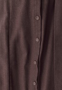 Hnědá látková košile s knoflíkovým zapínáním, obsahující pět kulatých knoflíků a hladkou texturu. Látka vypadá jako lehká a jemně se skládá.