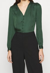 Blusa di satin verde con scollatura a V, bottoni frontali e maniche lunghe a sbuffo, abbinata a pantaloni neri a vita alta. Tessuto liscio.