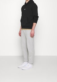 Felpa nera con cappuccio e tasca a marsupio, indossata sopra una maglietta beige. I pantaloni grigi e le sneakers bianche completano l'outfit.