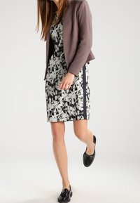 Černobílé šaty s vzorem a přiléhavým střihem, doplněné o texturovanou hnědou blazer a černé boty.