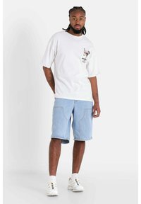 Vit bomulls-t-shirt med grafiskt tryck fram, ihop med ljusblå denimshorts. Modellen bär vita sneakers och ankelstrumpor, stående mot en neutral bakgrund.