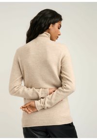 Kobieta z długimi ciemnymi włosami, ubrana w beżowy sweter z golfem i czarne spodnie, stojąca z rękami skrzyżowanymi za plecami.