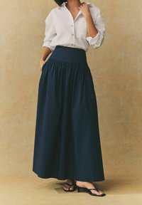 Jupe maxi bleu marine avec une taille haute et un design froncé. Associée à une chemise blanche à boutons. Le tissu texturé met en valeur la silhouette.
